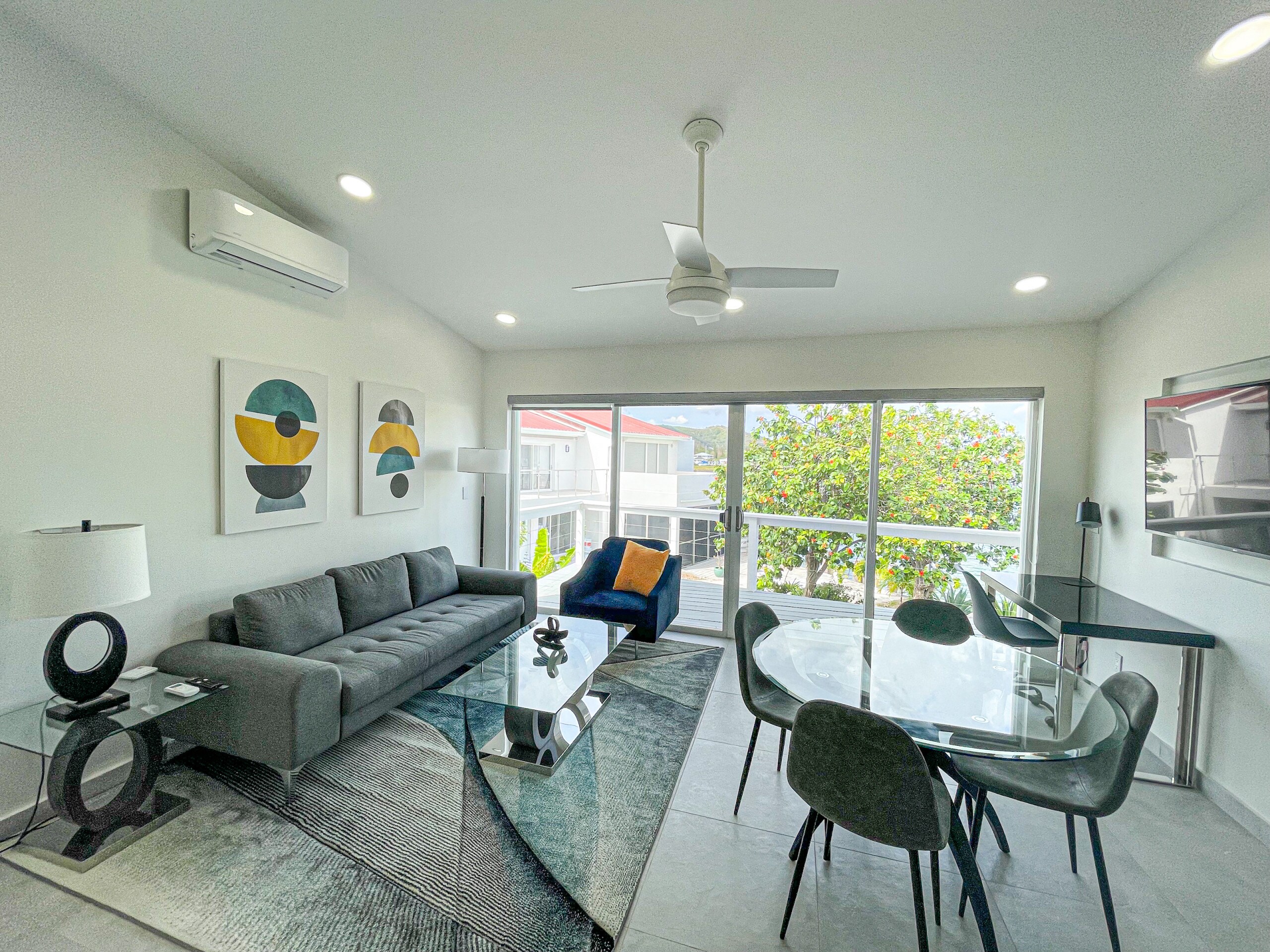 Stylish and Spacious one-Bedroom Apartment on the waterfront with an open plan interior, a round glass dining table, comfy grey sofa, a wall-mounted flat-screen TV, workspace area, tall glass walls, sliding doors, and a private balcony at Jolly Harbour Villa Rentals