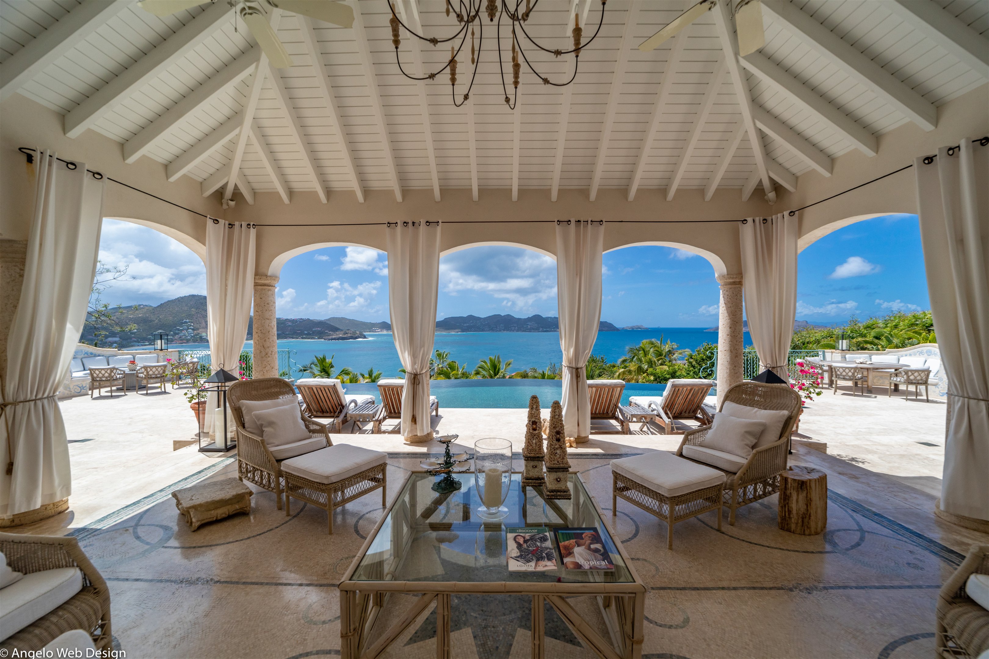 Beautiful opened living room, with woven rattan sofas, white cotton cushions, columns & long white drapes, mosaic tiling. Reminiscent of a Roman theater. Panoramic views. 