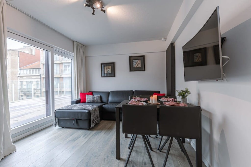 Modern and cozy apartment featuring a spacious living area with a comfortable sofa, dining table for four, and a flat-screen TV. Large windows offer natural light, and stylish decor enhances the welcoming atmosphere for a relaxing stay.