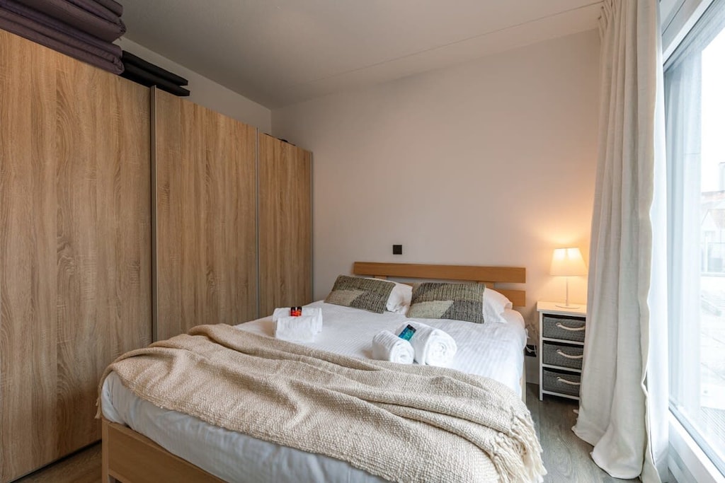 Cozy bedroom with a comfortable double bed, soft linens, and plush pillows for a restful sleep. The room features ample storage with a large wardrobe and bedside table with a lamp. Natural light fills the space through the window, creating a peaceful
