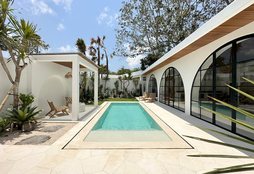 Large and sunny outdoor area with a white terrazzo swimming pool, two day beds, and a shaded outdoor lounge