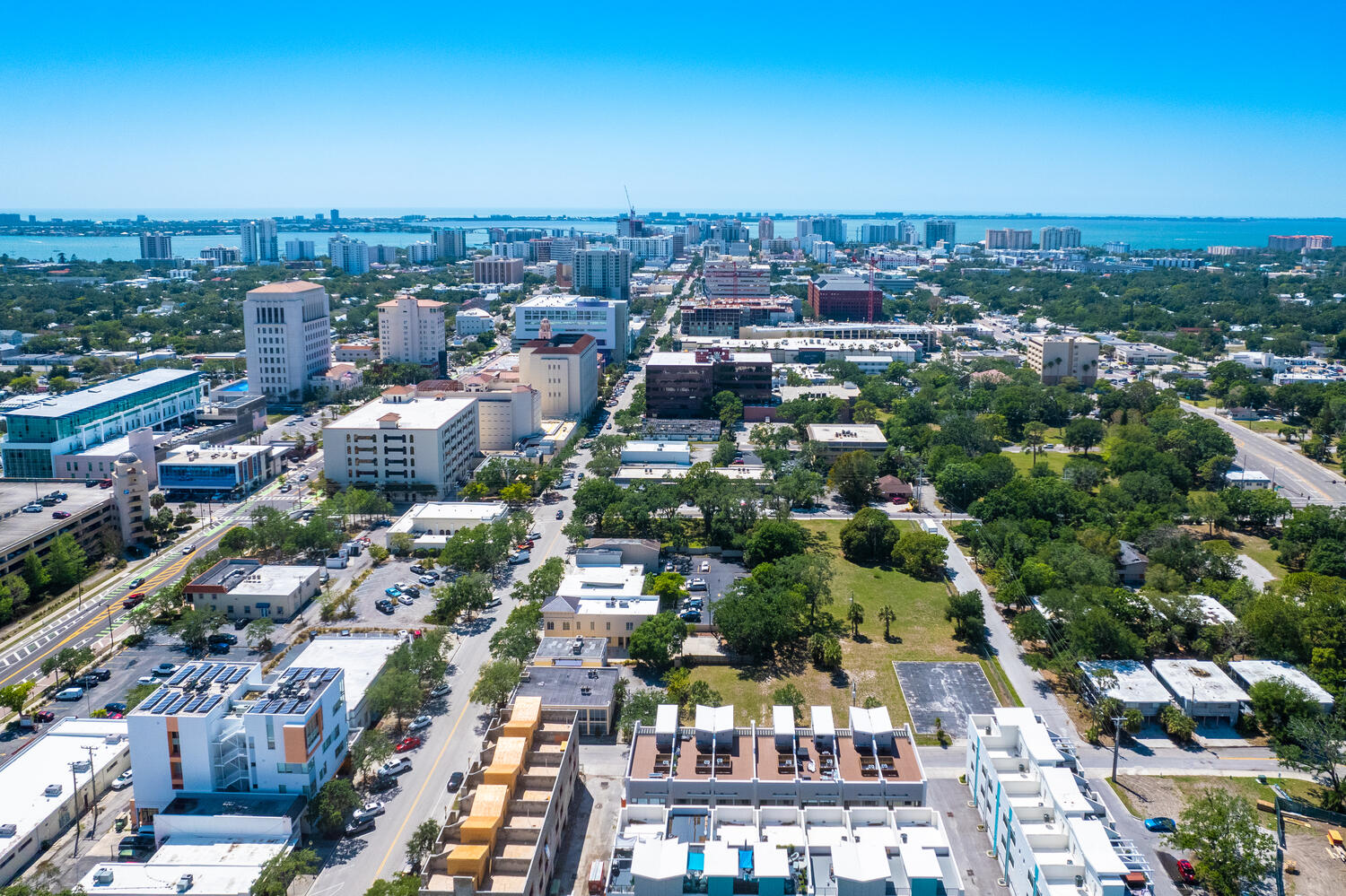 Aerial view of a cityscape with various buildings, trees, and roads leading to a distant waterfront under a clear blue sky.