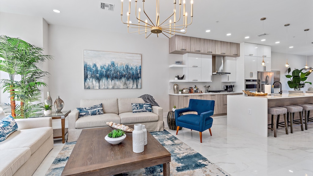 Modern living room and kitchen with white walls, hanging light fixture, blue and beige furniture, a large plant, abstract wall art, and a wooden coffee table with decorative items.