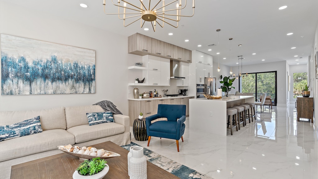 Modern open-concept living room and kitchen with white cabinetry, a beige sofa, blue armchair, island with bar stools, and decorative lighting. A large abstract painting hangs above the sofa.