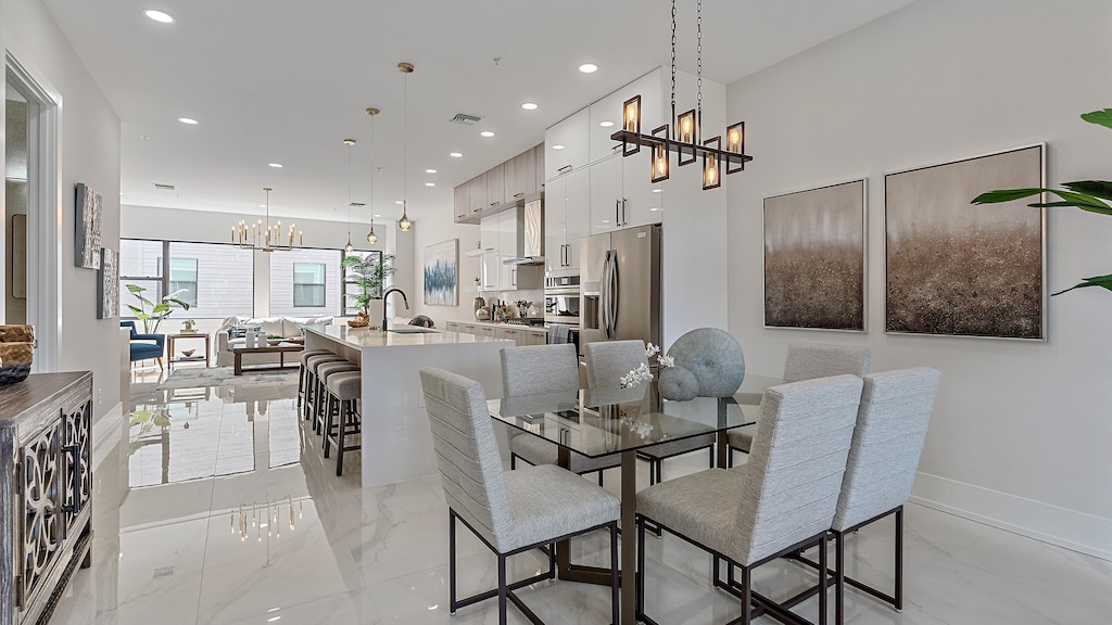 Modern open-concept kitchen and dining area with light-colored walls and floor, glass dining table with cushioned chairs, bar stools at the kitchen island, and contemporary light fixtures.