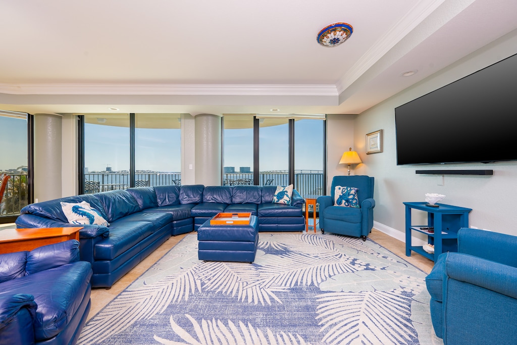 The spacious living room features a large blue sectional sofa, coastal decor, and an expansive balcony view of the bay