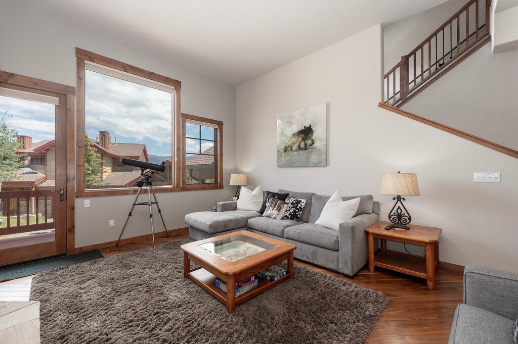 - Bright and spacious living room with large windows and cozy gray furnishings - Telescope offers a fun way to stargaze or take in mountain and rooftop views - Soft rug, wood tables, and stairway access complete the inviting layout