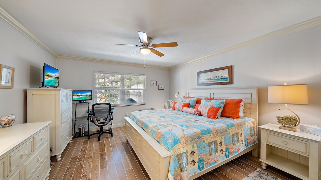A bedroom with a bed covered in a colorful quilt, two bedside tables, a ceiling fan, a dresser with a TV, and a desk with a chair and computer setup near a window with blinds.