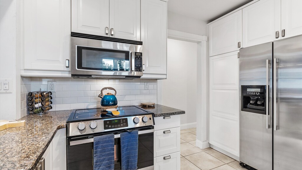 A modern kitchen with white cabinets, stainless steel appliances, and a countertop stove. A blue kettle sits on the stove, and there are towels hanging on the oven door.