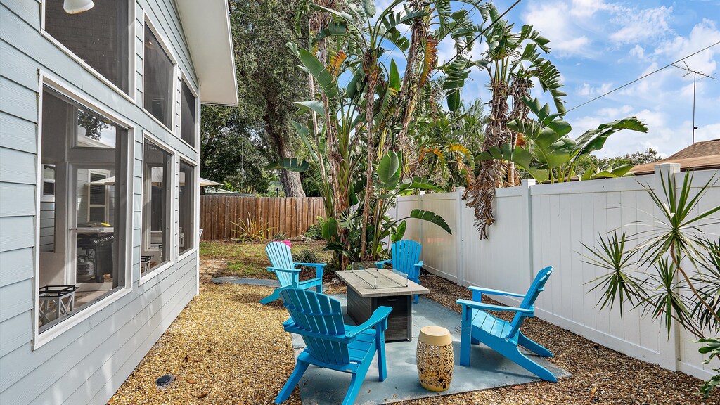 Outdoor seating area with a rectangular table and four blue chairs. The area is surrounded by a white fence and lush green plants. A nearby house has large windows overlooking the space.