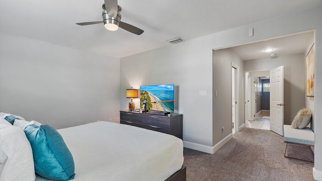 A bedroom with gray walls and carpet features a bed with white linens and blue pillows, a dresser with a TV, a ceiling fan, a side table with a lamp, and an open door leading to a bathroom.