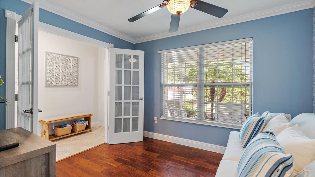 A room with blue walls features a white couch with striped pillows, a ceiling fan, a large window with blinds, and a glass-paned door opening to a hallway with a wooden bench.