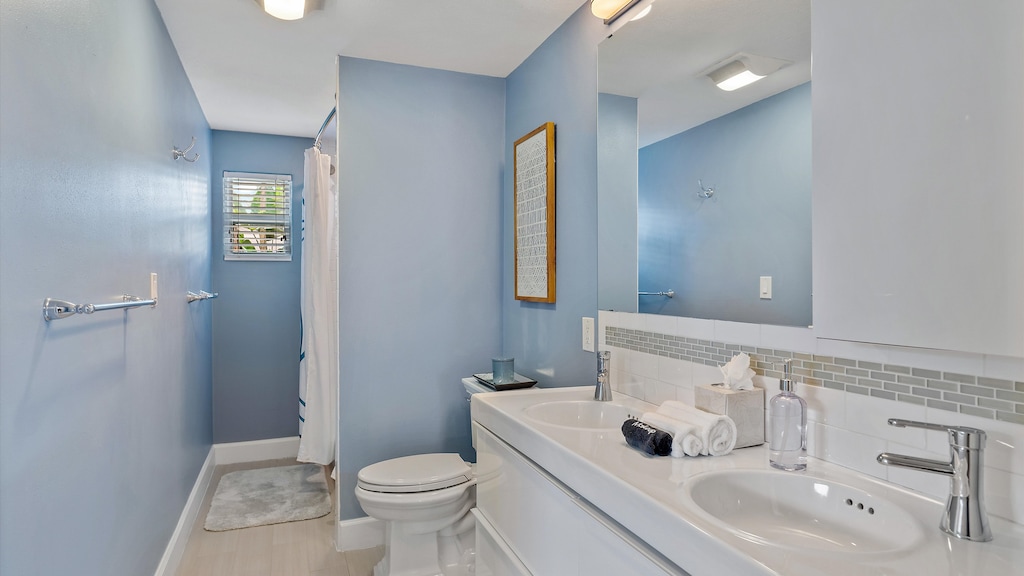 A modern bathroom with blue walls, a double sink vanity with a large mirror, a toilet, a shower with a white curtain, and a small window providing natural light.