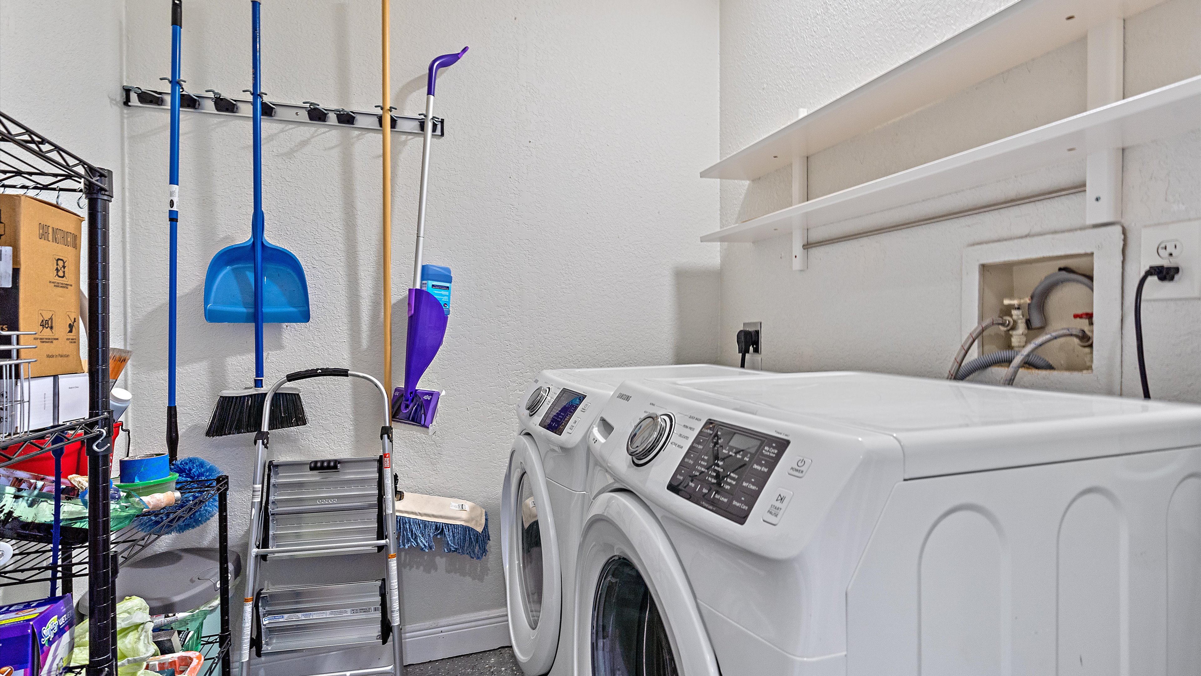 A laundry room with a white washer and dryer, cleaning supplies, a ladder, and storage shelves mounted on the wall.