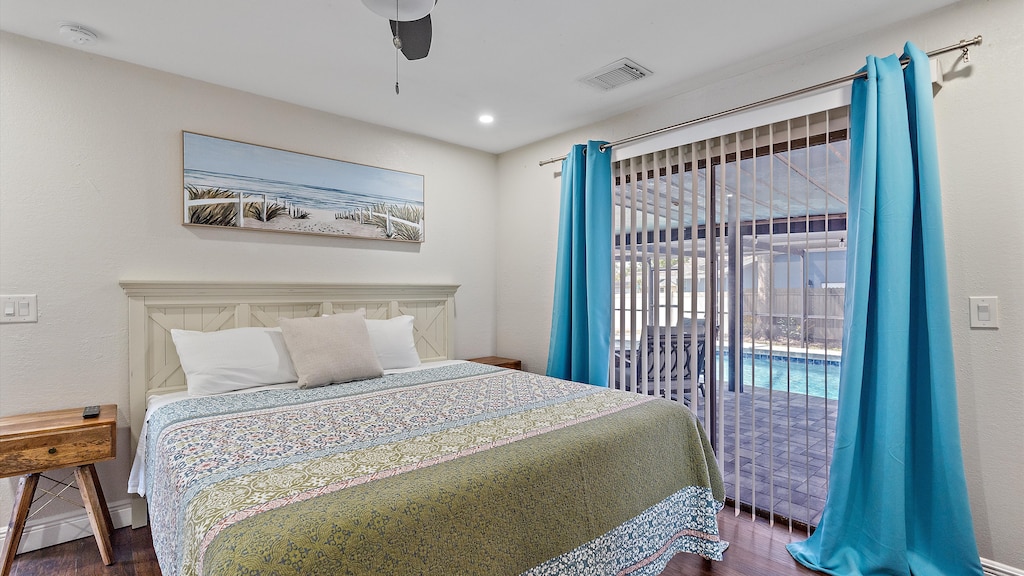 A bedroom with a large bed featuring a colorful quilt, two nightstands, and blue curtains covering sliding glass doors that open to a view of a backyard pool. A landscape painting hangs on the wall above the bed.