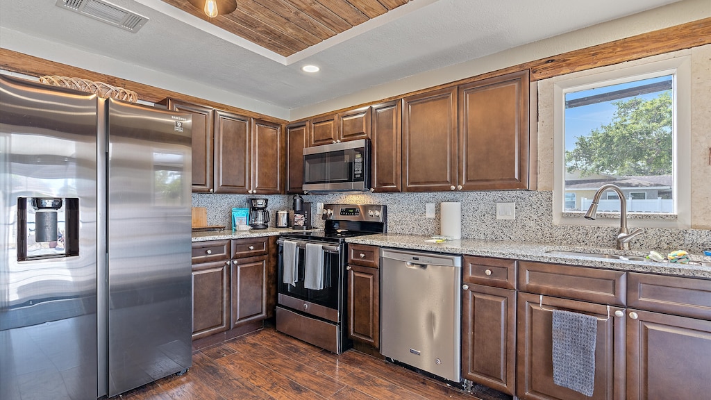 A modern kitchen with dark wooden cabinets, stainless steel appliances including a refrigerator, oven, microwave, and dishwasher, a granite countertop, and a window above the sink.