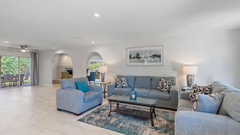 A modern living room with light gray sofas, a blue armchair, glass coffee tables, and decorative pillows. There's a large painting on the wall, two table lamps, and a dining area visible in the background.