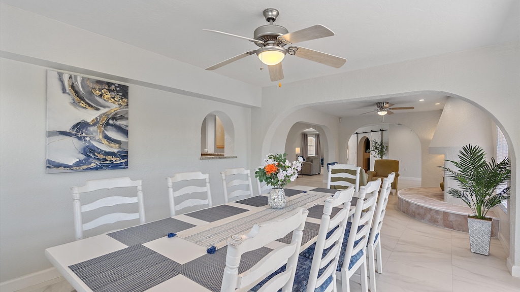 A dining room with a long table and white chairs, a ceiling fan, an abstract painting on the wall, and a view into a living area with a fireplace and another ceiling fan.
