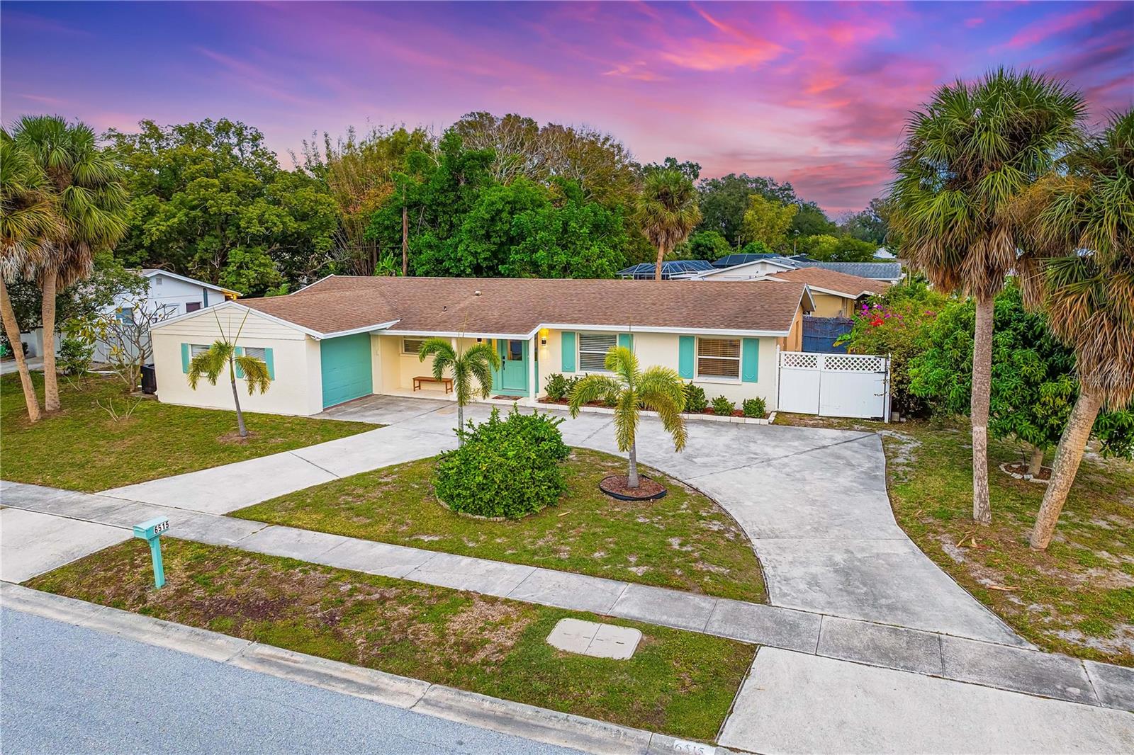 A single-story house with a curved driveway, palm trees, and a well-maintained yard under a vibrant, purple and pink sunset sky.