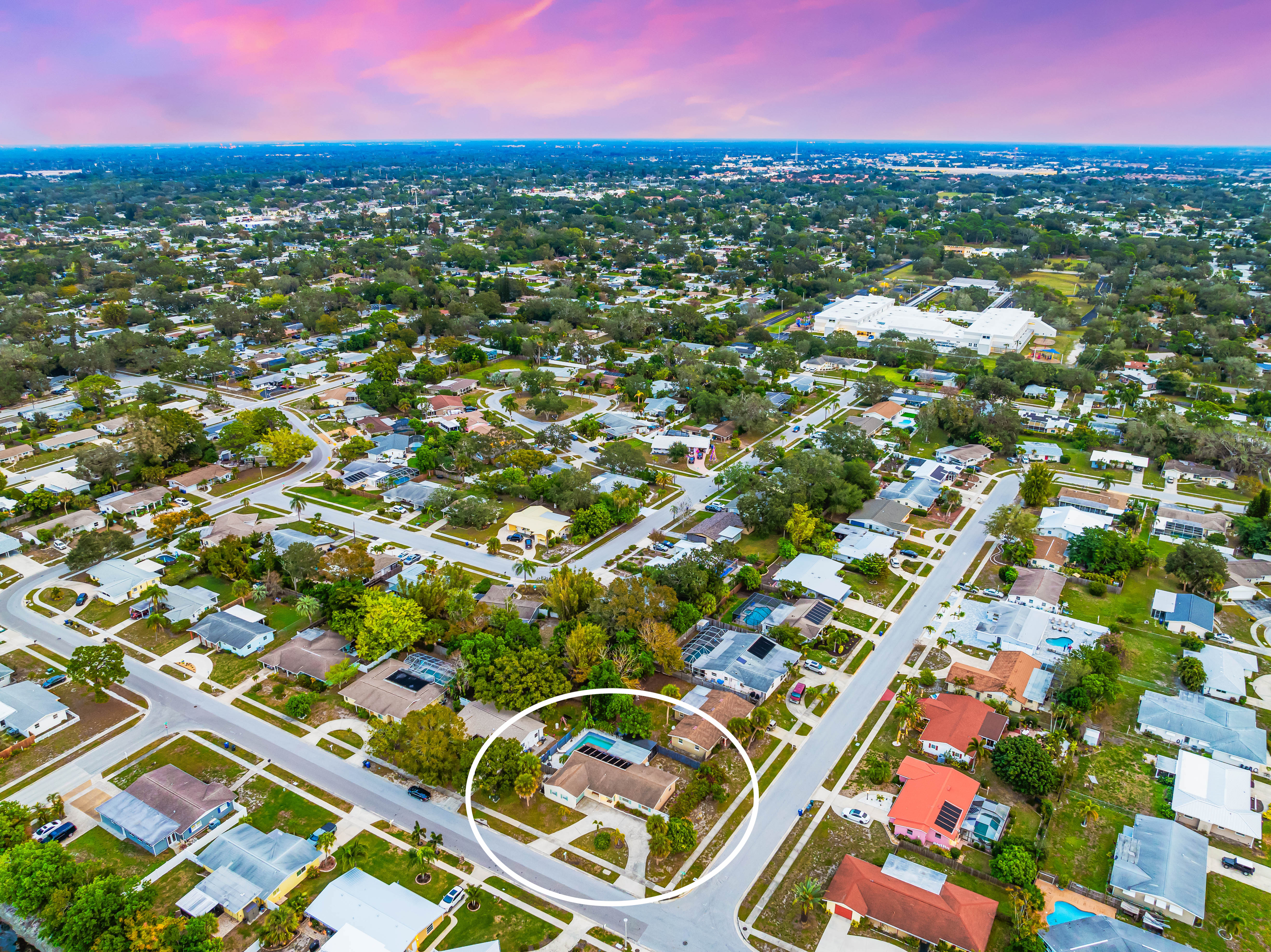 Aerial view of a residential neighborhood during sunset, featuring multiple houses and tree-lined streets. One house is circled, highlighting its location.