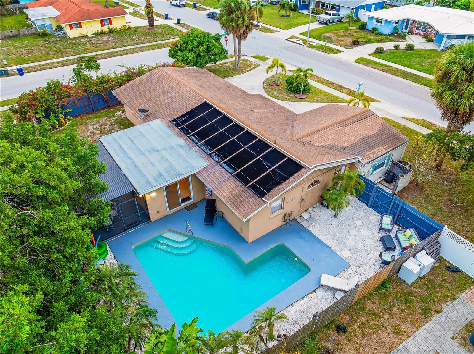 Aerial view of a house with a beige roof featuring a swimming pool in the backyard, partially covered by a screen enclosure, surrounded by other houses and trees on a sunny day.