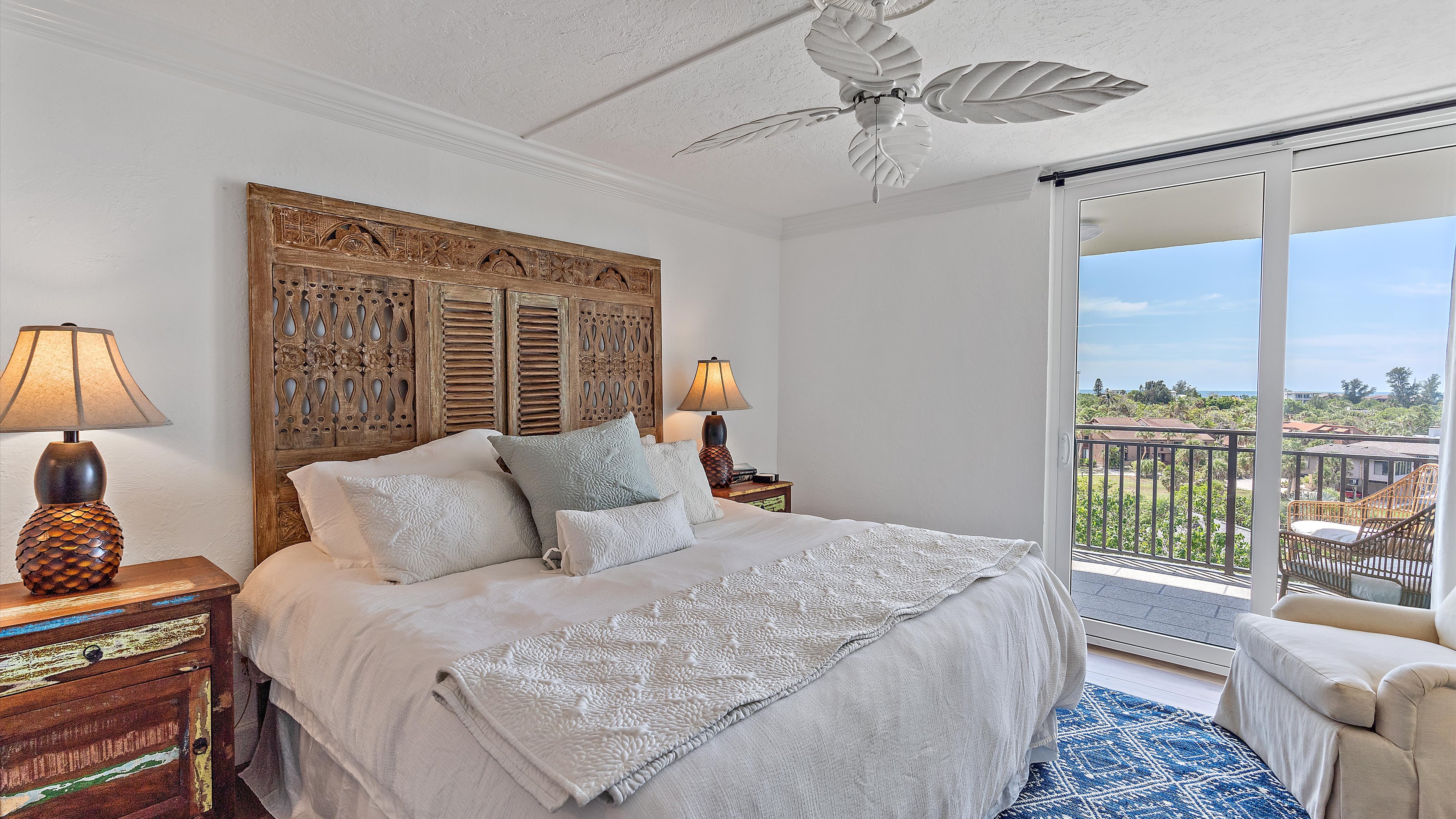 A bedroom with a large bed features a wooden headboard, two bedside tables with lamps, a ceiling fan, and glass doors opening to a balcony with outdoor furniture and a view of trees and buildings.
