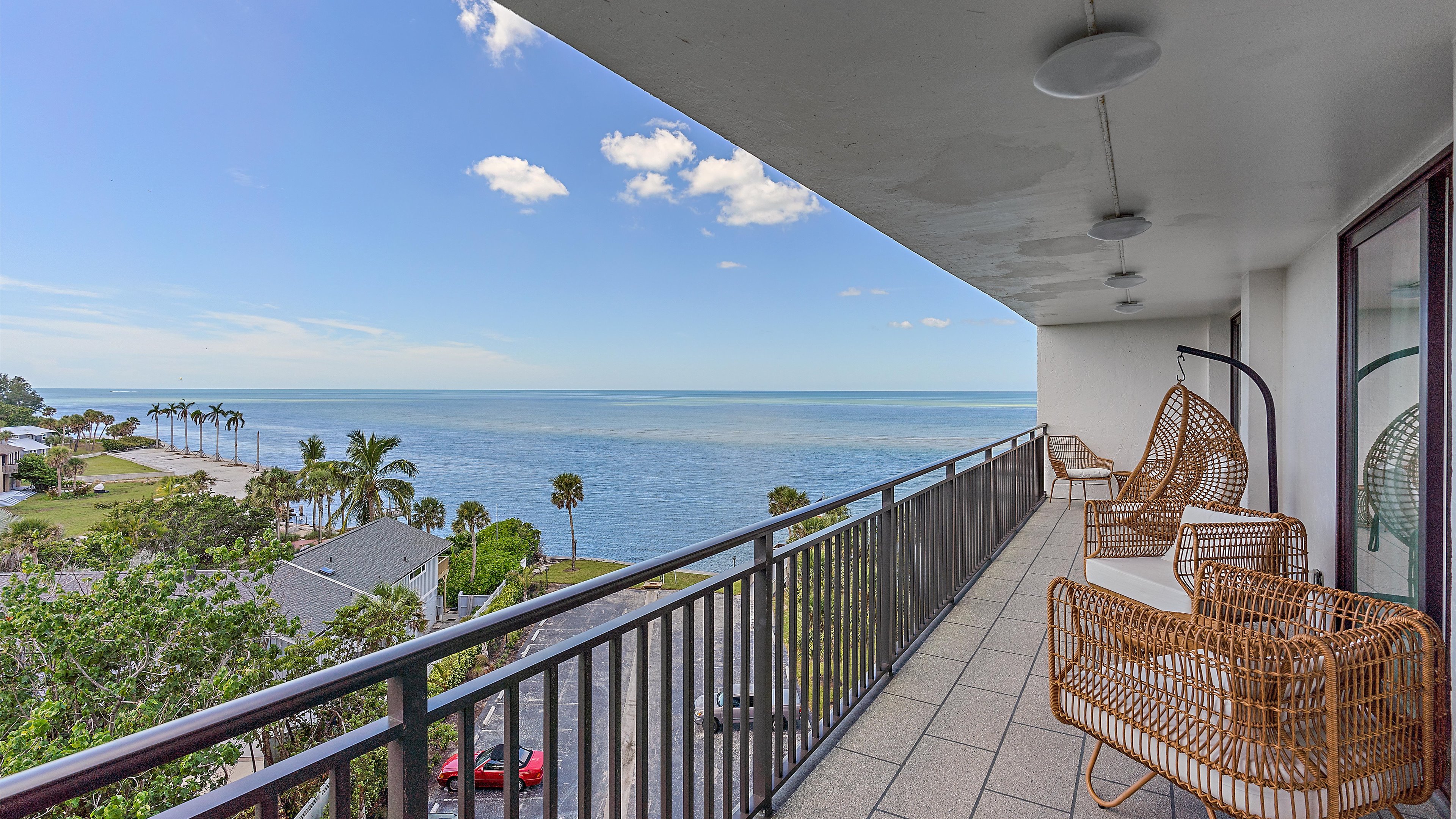 A large balcony with wicker chairs overlooks a calm ocean, a sandy beach with palm trees, and a clear blue sky with a few clouds.