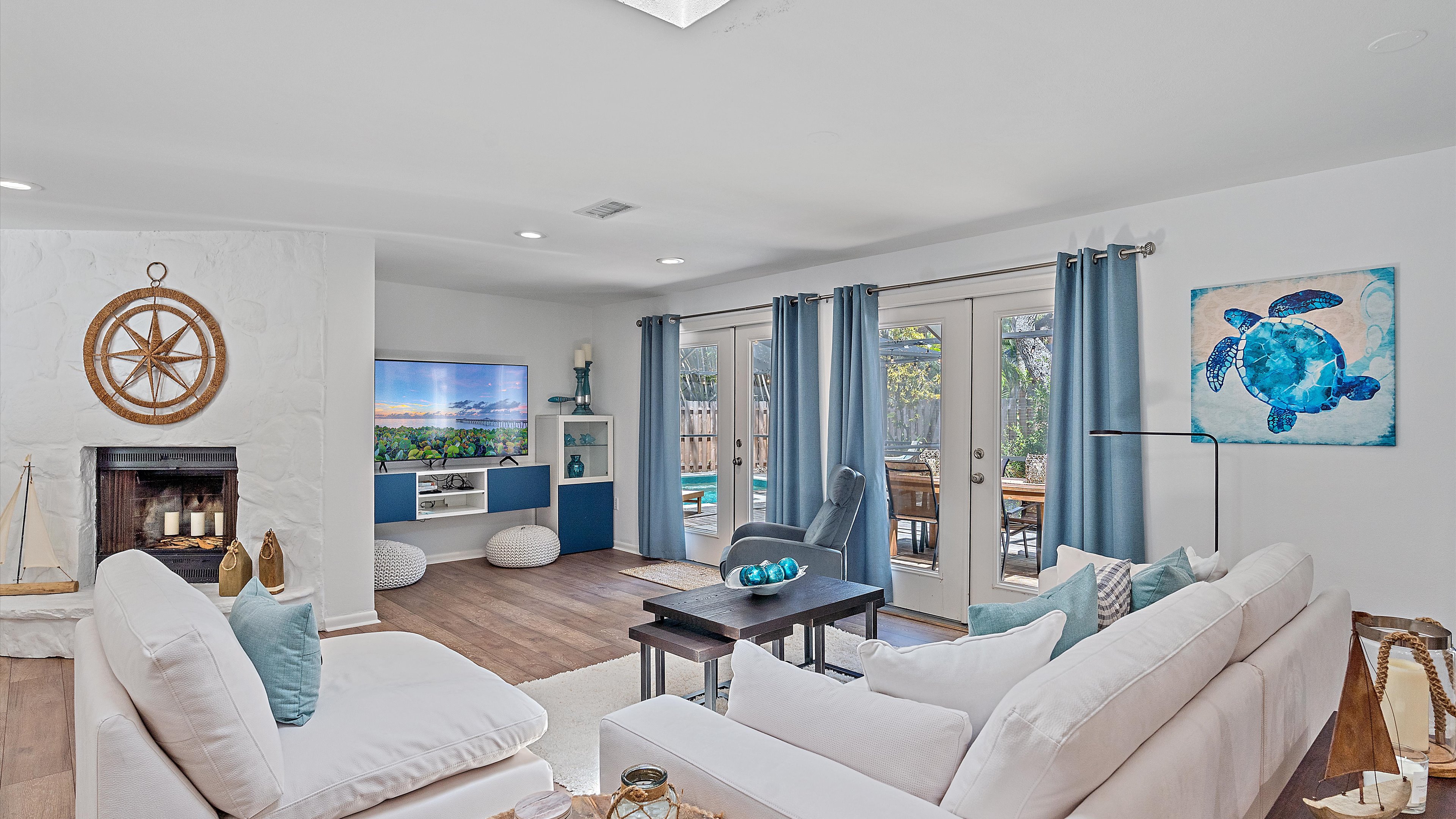 A bright living room with white couches, a wall-mounted TV, nautical decor, and a sliding glass door leading to an outdoor patio with a view of a pool. Blue and white color scheme is predominant.