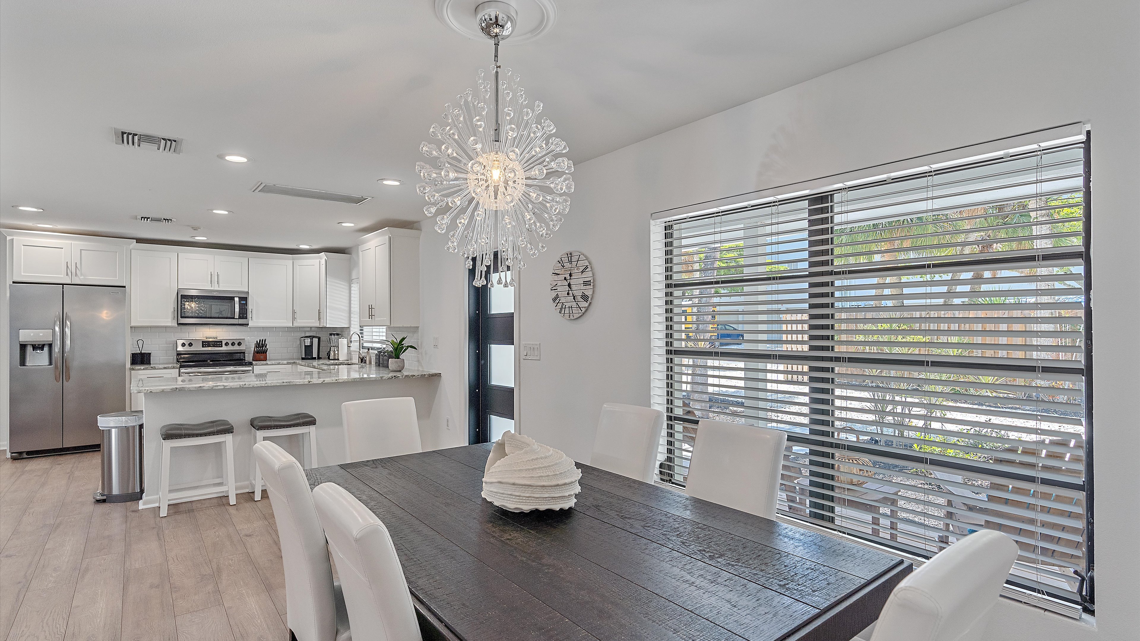 A modern kitchen with a large wooden dining table and white chairs. The kitchen features stainless steel appliances, white cabinetry, and a decorative light fixture. Light streams through window blinds.