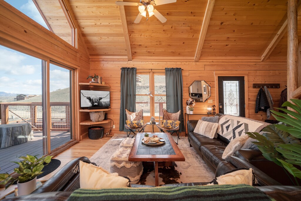 A cozy cabin living room with wooden walls and ceilings, large windows, and modern furnishings, including leather sofa, a TV mounted on the wall.