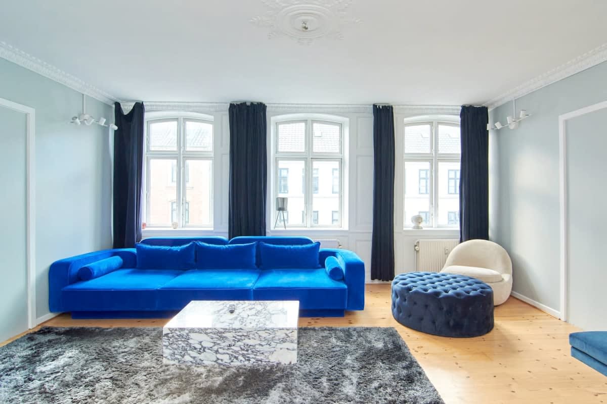 This gorgeous living room boasts light blue walls that create a calming and soothing atmosphere. As you step in, you'll be greeted by a luxurious blue velvet sofa and matching pouf that invites you to sit back and relax.