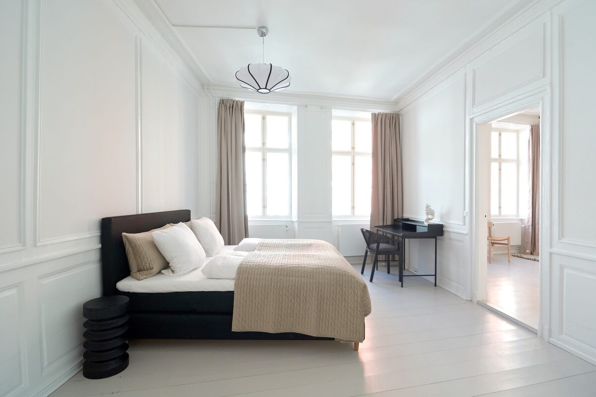The apartment features two bright and comfortable bedrooms with elegant white walls, white floors, high ceilings, and beautiful beige curtains that create a cozy and inviting atmosphere.