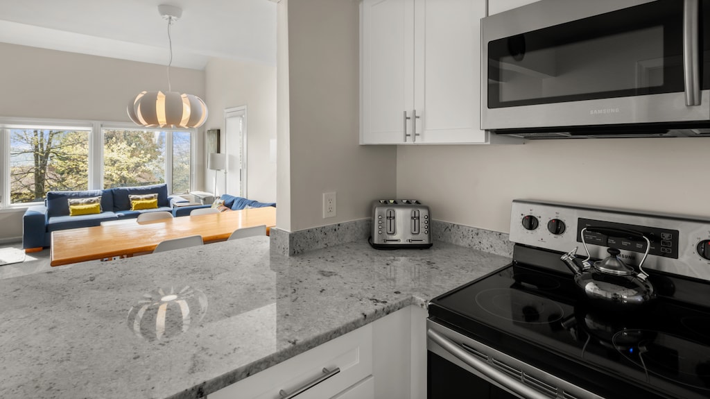 A modern kitchen with gray countertops, a stainless steel stove, a microwave, a toaster, and a teapot. The background shows a dining area with a wooden table, chairs, and a large window.