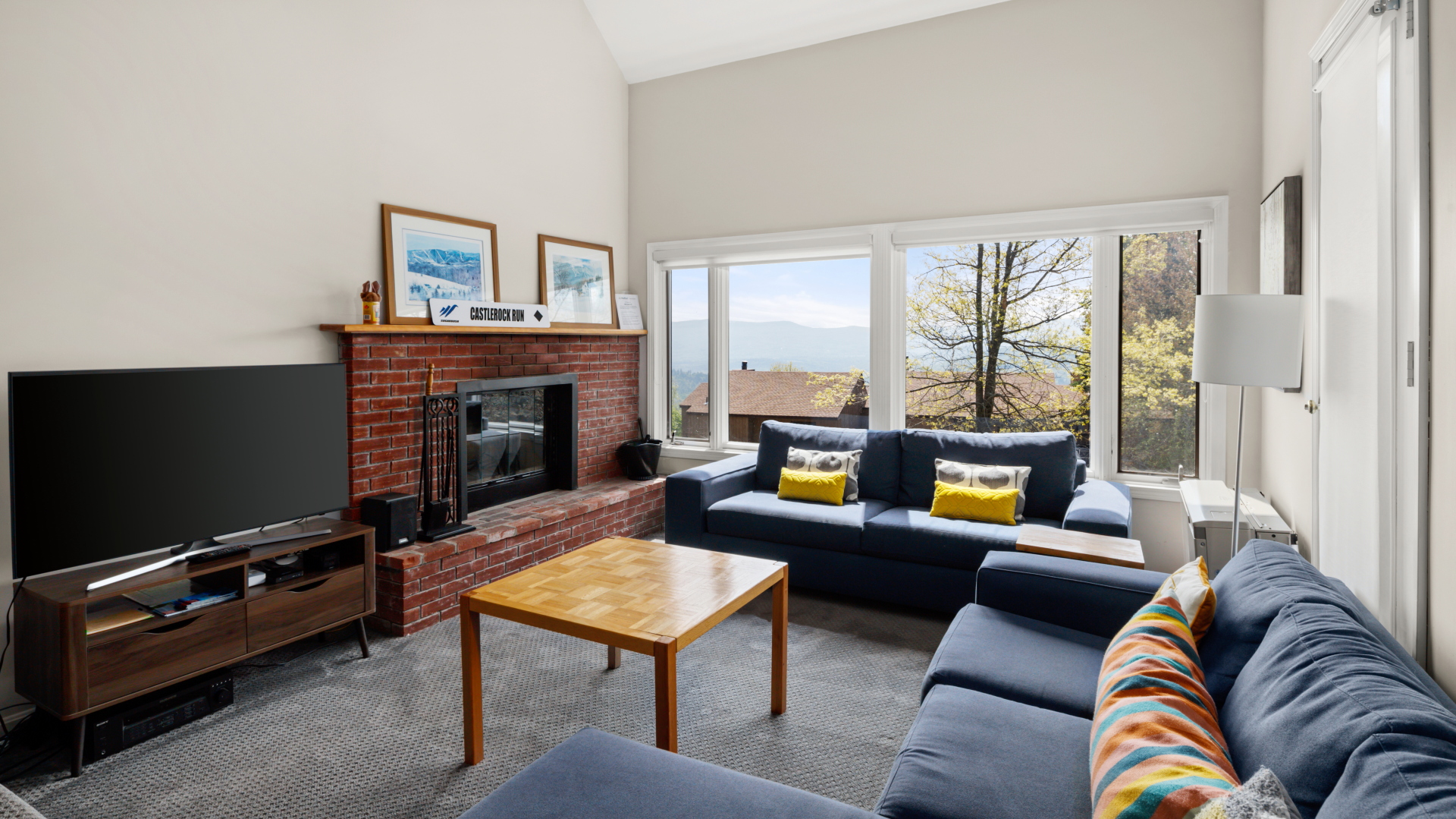 A living room with two blue couches, a wooden coffee table, a brick fireplace with a TV, and large windows offering a view of trees and distant water. Framed pictures and a clock adorn the mantel.