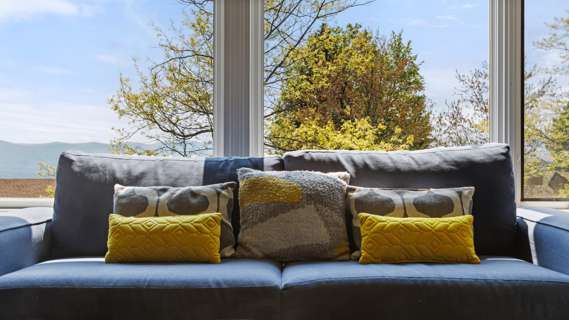 A blue couch with decorative pillows is positioned in front of large windows, showcasing a scenic view of trees and mountains outside.