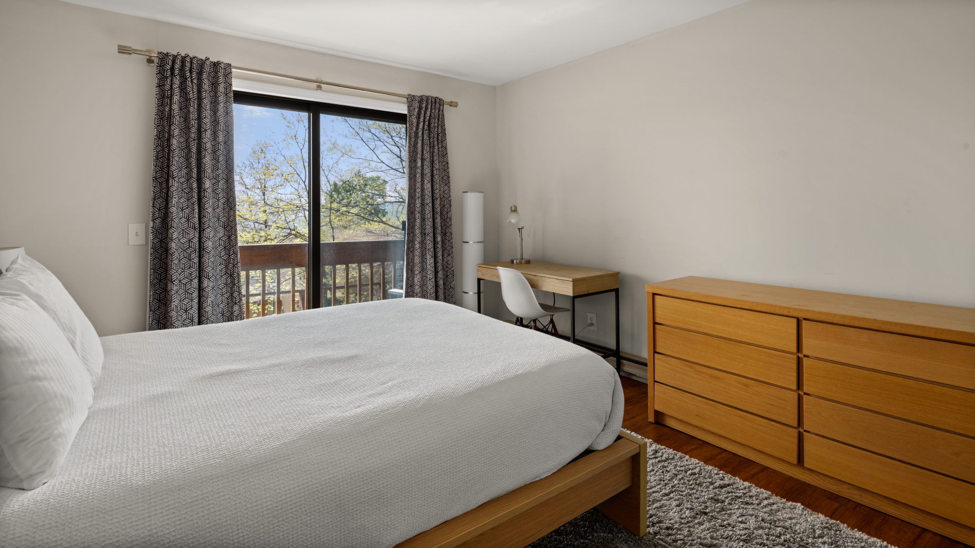 A bedroom with a bed, wooden dresser, and a small desk with a chair near a sliding glass door that leads to a balcony. The room has white walls and curtained windows.