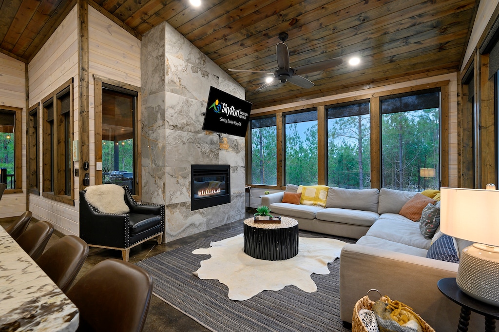 A modern living room with a fireplace, large sectional sofa, and flat-screen TV. The room features wooden walls, large windows, and a cowhide rug.