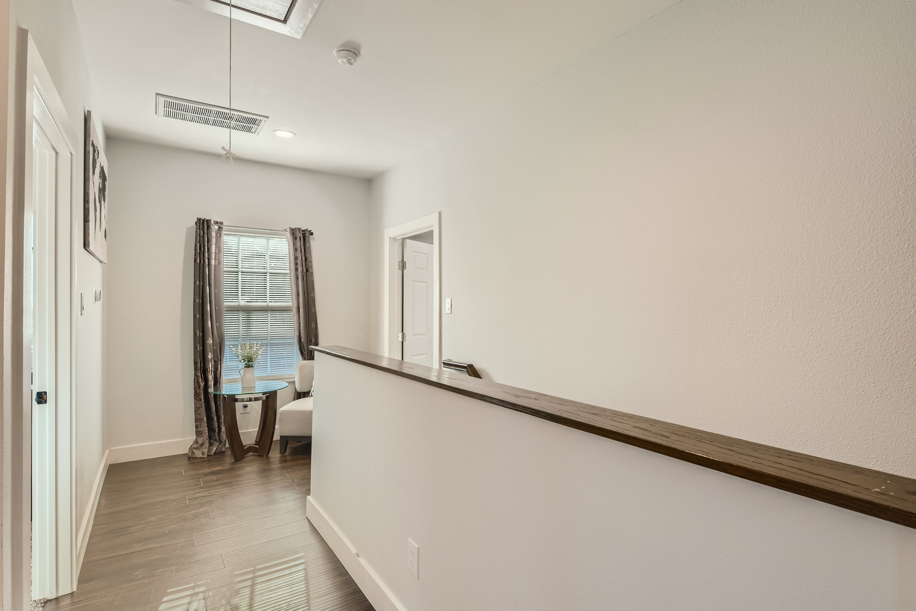 A hallway with a wooden railing on one side leads to a window with curtains. A small table with a vase and chairs is placed near the window. The floor is wooden, and the walls are painted white.