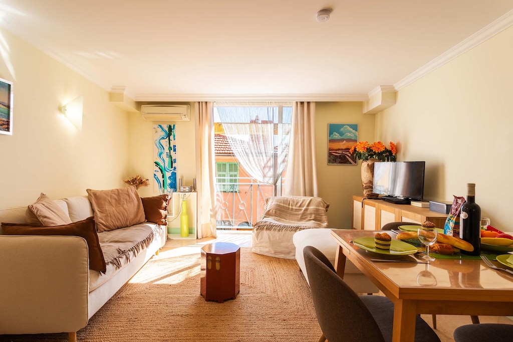 Bright living room with a comfortable sofa, TV, dining table, and window offering a view of the sunny outdoors. Perfect for relaxing.