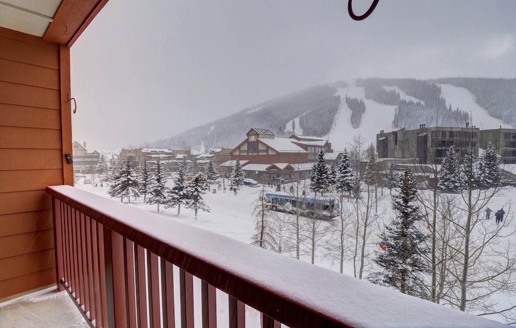 Snowy mountain village viewed from a balcony, with buildings, evergreen trees, and a bus in the snow-covered landscape. Ski slopes are visible in the background under a cloudy sky.