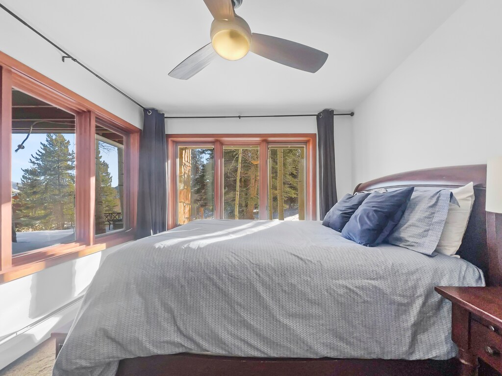 A bedroom with a large bed, wood frame windows showing a forest view, dark curtains, a ceiling fan, and wooden nightstands.
