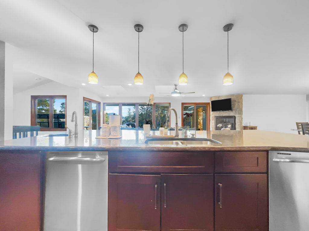 A modern kitchen with a large island sink, pendant lights above, stainless steel dishwasher, and wooden cabinets. Two sets of double doors open to a deck with seating. A fireplace is visible in the background.