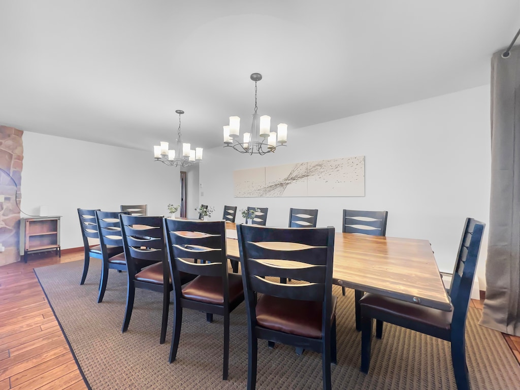 A spacious dining room with a large rectangular wooden table surrounded by black chairs. Two chandeliers hang from the ceiling. The room has wooden floors and a large wall painting.