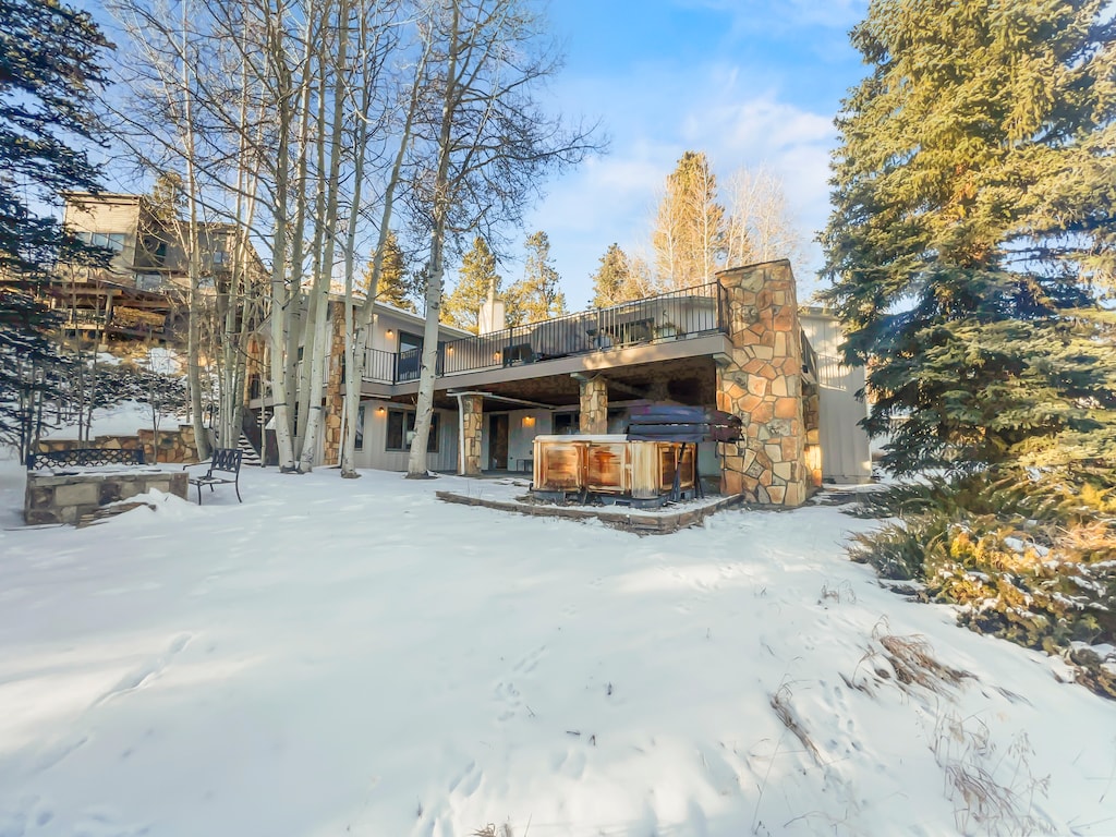 A modern two-story house with a stone facade, surrounded by trees and snow. The backyard features an outdoor hot tub on a patio and a fire pit area.
