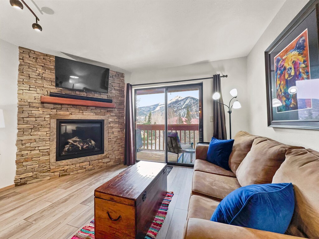 A cozy living room with a stone fireplace, wall-mounted TV, brown sofa, and wooden chest. A sliding glass door opens to a balcony with mountain views. A vibrant painting and floor lamp decorate the space.
