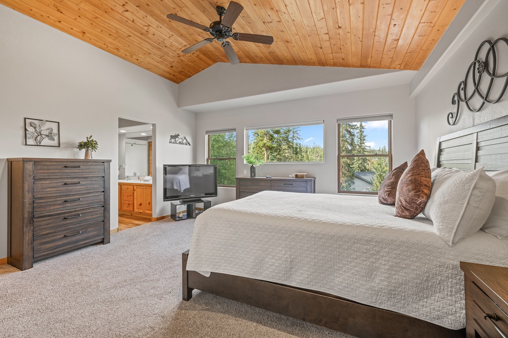 Spacious primary bedroom featuring a striking vaulted wood-paneled ceiling with a ceiling fan, plush carpeting, and large windows offering tranquil forest views. Includes an en-suite bathroom and a cozy seating area with a Smart TV, perfect for a relaxing