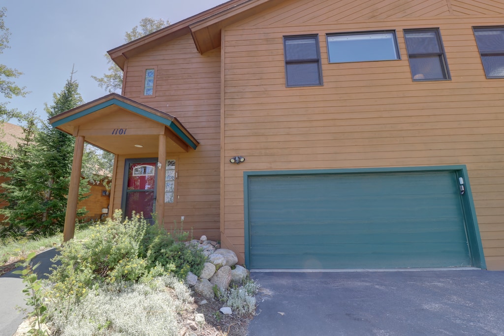 A two-story wooden house with a green garage door, a red front door, and surrounding greenery. The house number "1101" is displayed above the front entrance.