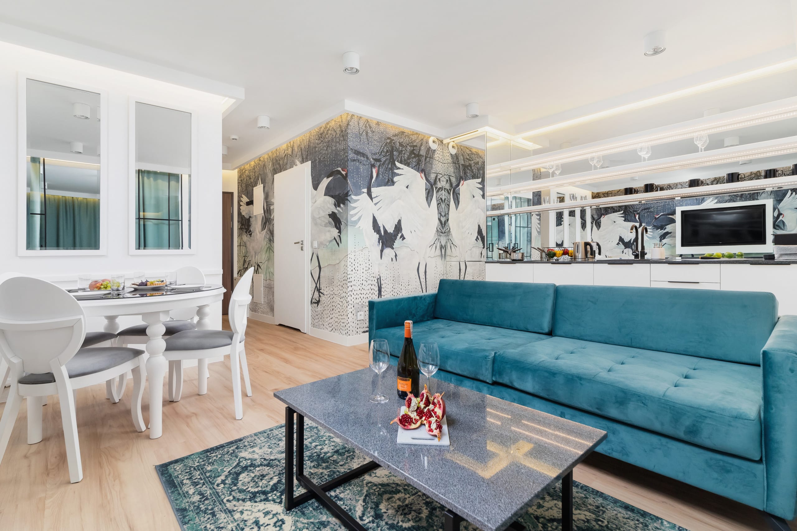  A stylish living area featuring a turquoise sofa, artistic wallpaper, a flat-screen TV, and a coffee table. The room is designed with modern elegance.