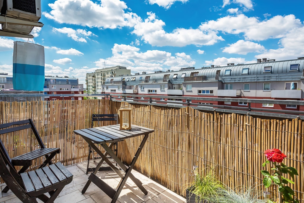 A spacious balcony with a wooden floor, comfortable outdoor furniture, and a panoramic view of the city under a bright blue sky with scattered clouds.