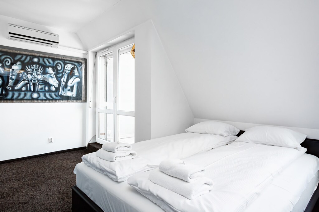 A bright bedroom with a double bed adorned with crisp white linens, slanted ceilings, and a large window allowing sunlight to flood the space.
